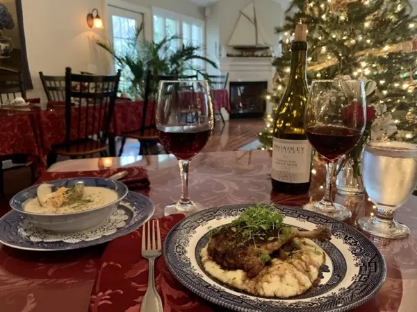 A holiday table with a plated entree, red wine and a Christmas tree behind